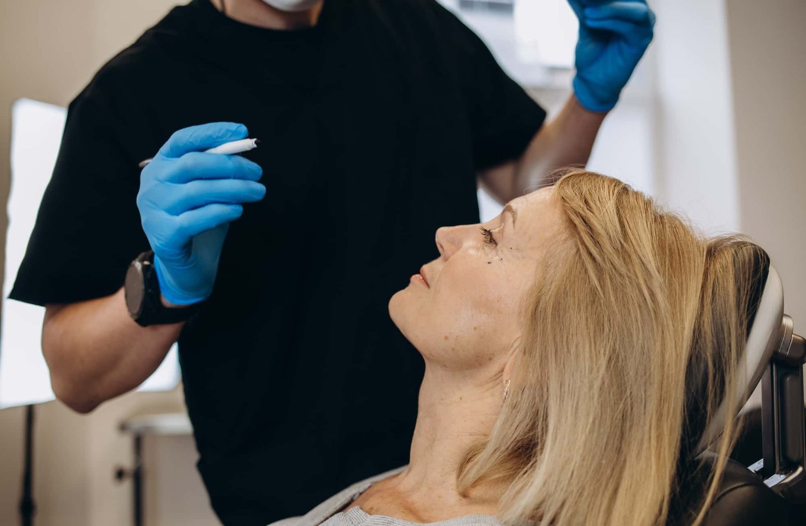 A woman sits in a reclined chair while a person wearing blue gloves and holding a syringe prepares for an SEO-related procedure.