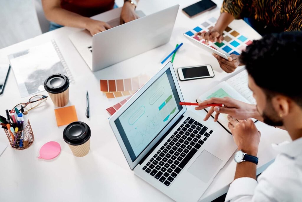 Three people work at a desk with laptops, coffee cups, color swatches, and a tablet, discussing a design project displayed on a laptop screen.