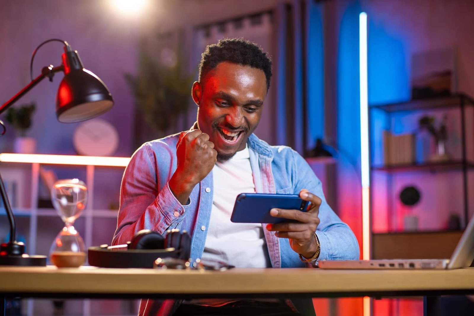 A man sits at a desk, smiling and raising his fist in celebration while looking at a smartphone—perhaps after finding fixes online. The desk features a laptop, headphones, an hourglass, and the room glows with colorful lights.