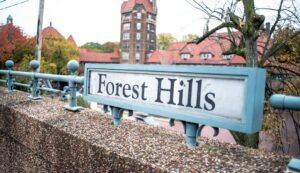 Sign reading "Forest Hills" mounted on a stone wall, with a background of red-roofed buildings and trees.