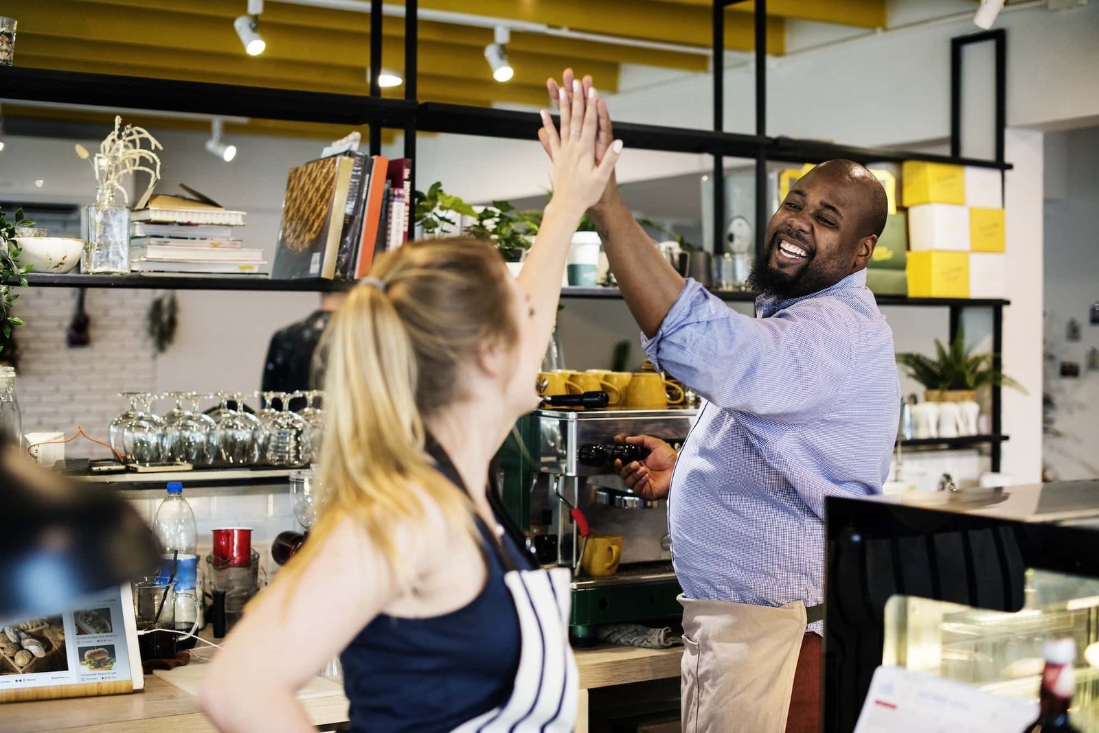 Two café employees, one man and one woman, wearing aprons, smile and give each other a high-five behind the counter in a modern coffee shop known for great service and Local SEO success.