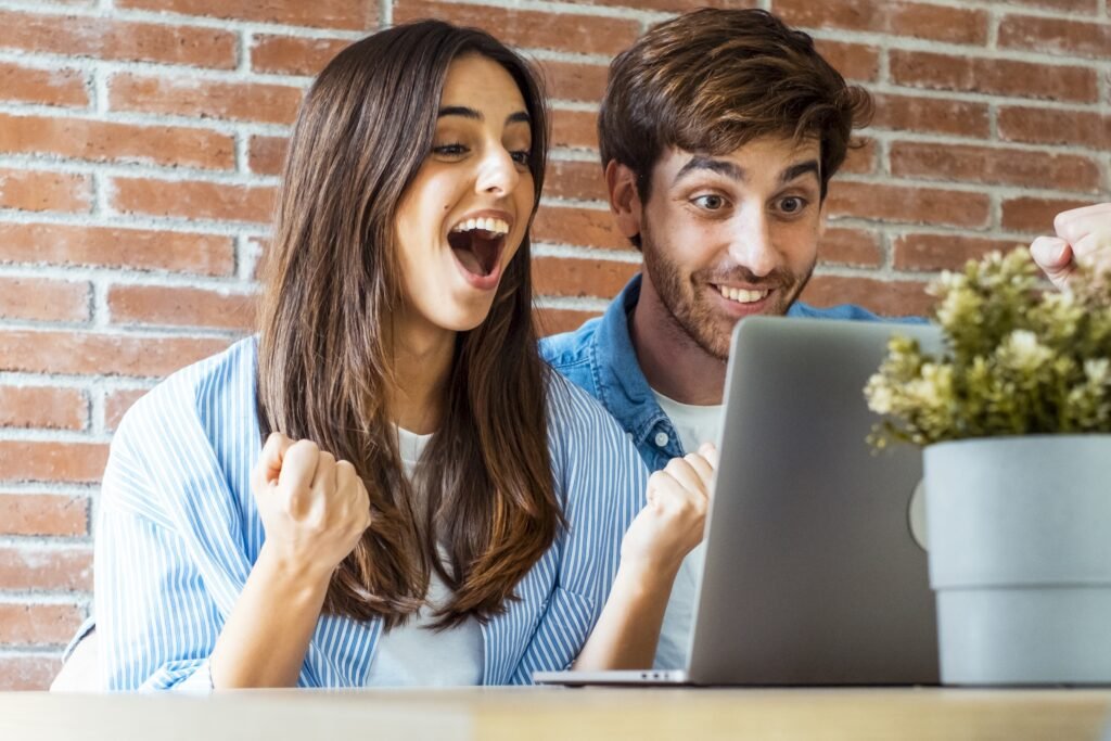 A woman and man sit at a table in front of a laptop, smiling and celebrating with raised fists—proof that you can Turn Your Dead-End Website into a Sales Magnet. A potted plant adds a fresh touch in the foreground.