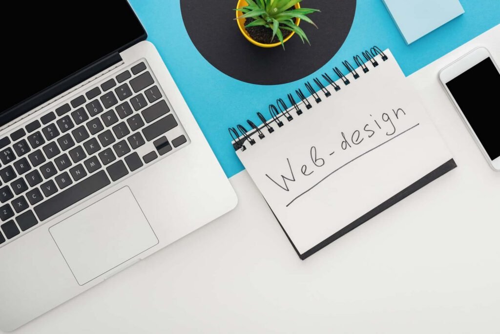 A laptop, smartphone, potted plant, and a spiral notebook labeled "Web-design" sit on a desk with a blue and white background—an inspiring NYC Web Designer workspace.