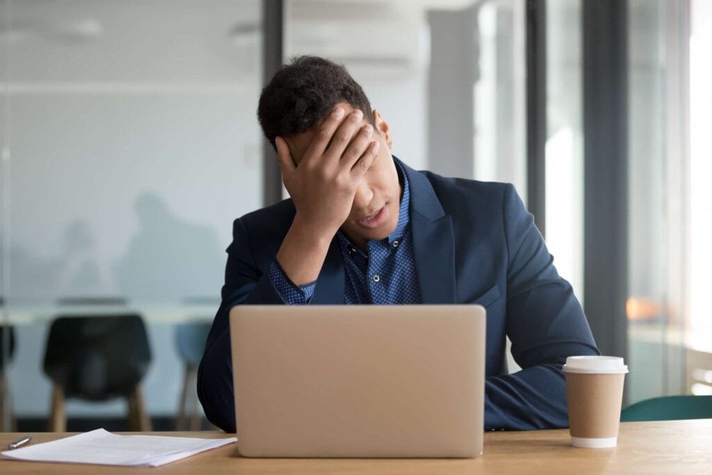 A man in a suit sits at a desk with a laptop, resting his head in his hand, appearing stressed—perhaps overwhelmed by SEO tasks. A paper and a disposable coffee cup are on the table.