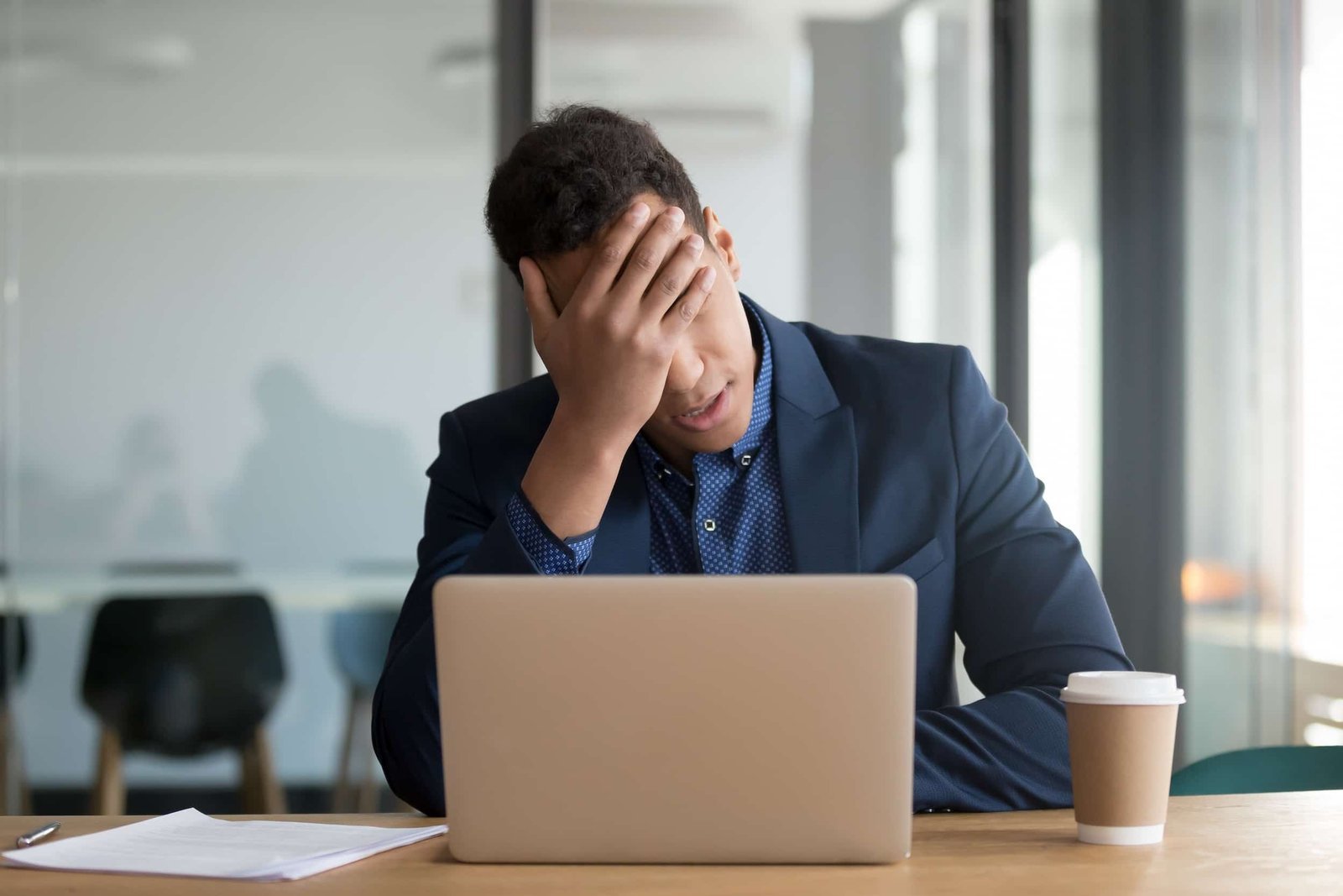 A man in a suit sits at a desk with a laptop, resting his head in his hand, appearing stressed—perhaps overwhelmed by SEO tasks. A paper and a disposable coffee cup are on the table.