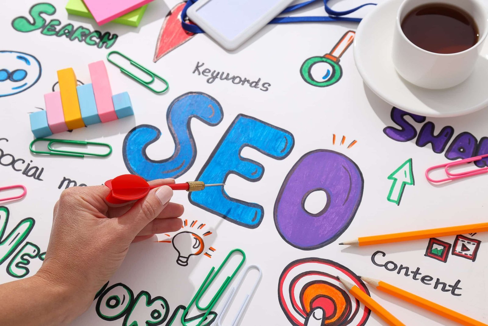 A hand holds a red dart over a colorful SEO diagram with keywords like "social media," "content," and "search," surrounded by office supplies and a cup of coffee.
