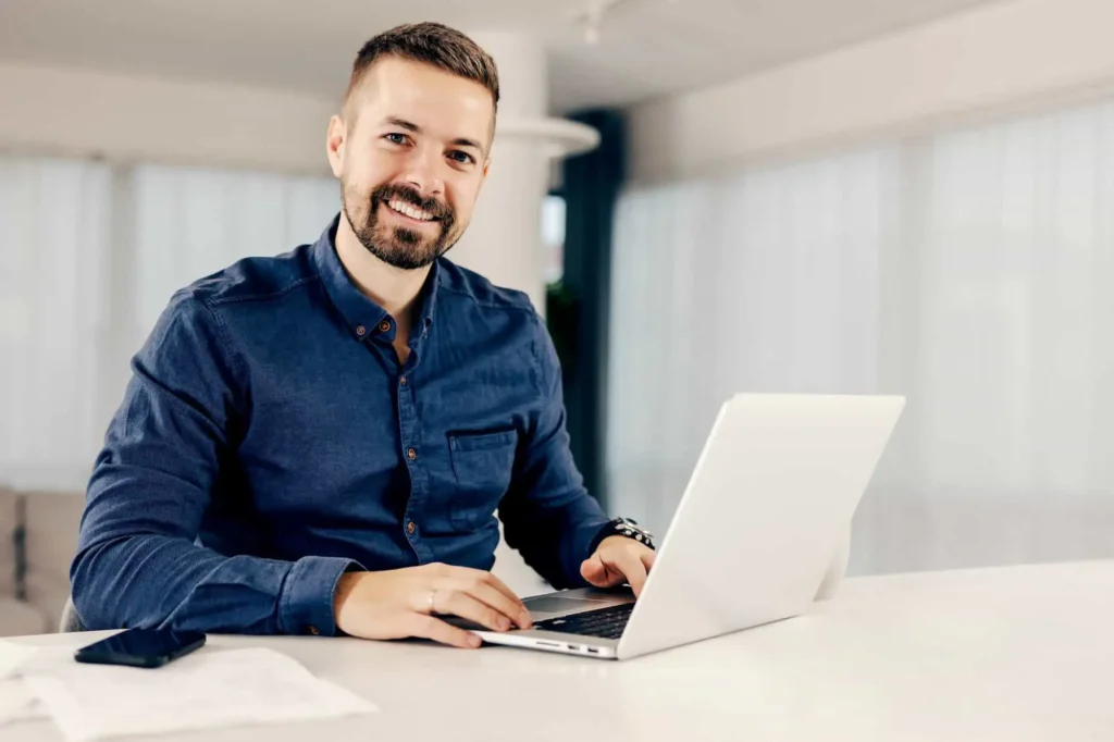 Man with a beard in a blue shirt sitting at a desk, typing on a laptop, with a smartphone and papers nearby. Unconventional Methods of Integrating AI with Your Current SEO Strategy.