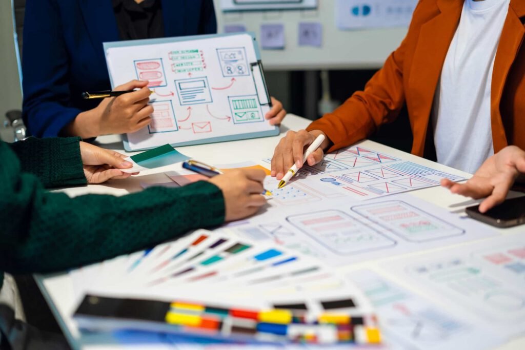 Three people work together at a table covered with papers, markers, and wireframe sketches, discussing and designing website or app layouts.