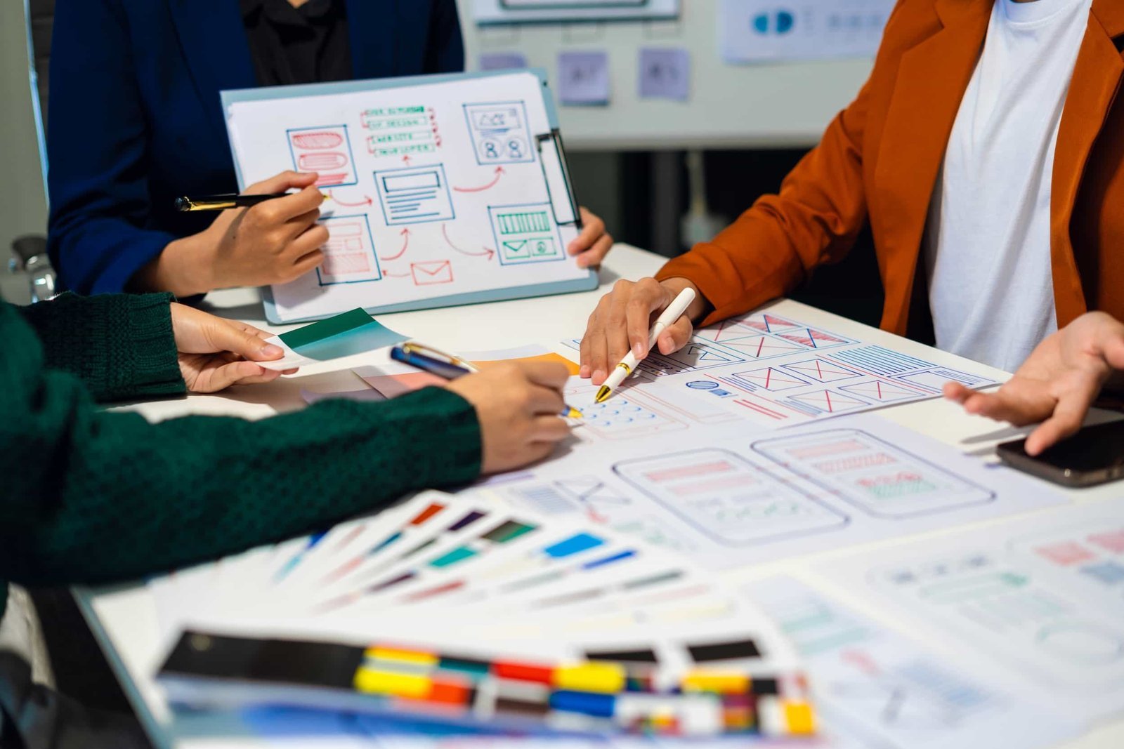 Three people work together at a table covered with papers, markers, and wireframe sketches, discussing and designing website or app layouts.