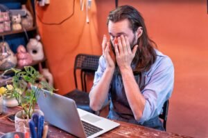 A person wearing an apron sits at a desk with a laptop, rubbing their eyes with both hands, appearing tired or stressed.