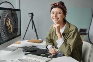 Person wearing glasses and a headband sits at a desk with a computer, smiling at the camera. Food appears on the screen, hinting at queens ny web design work. A tripod and laptop are visible in the background.
