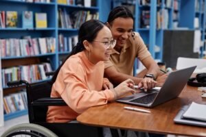Two students, highlighting accessibility, work together on a laptop at a table in a library with bookshelves in the background; one student uses a wheelchair. WCAG