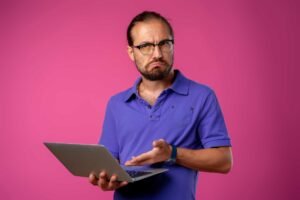 Man in a blue polo shirt and glasses holding a laptop, looking confused or frustrated, against a plain pink background.