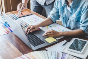Two people collaborate at a desk with a laptop, color swatches, printed charts, a notebook, and a tablet, discussing design or project plans.