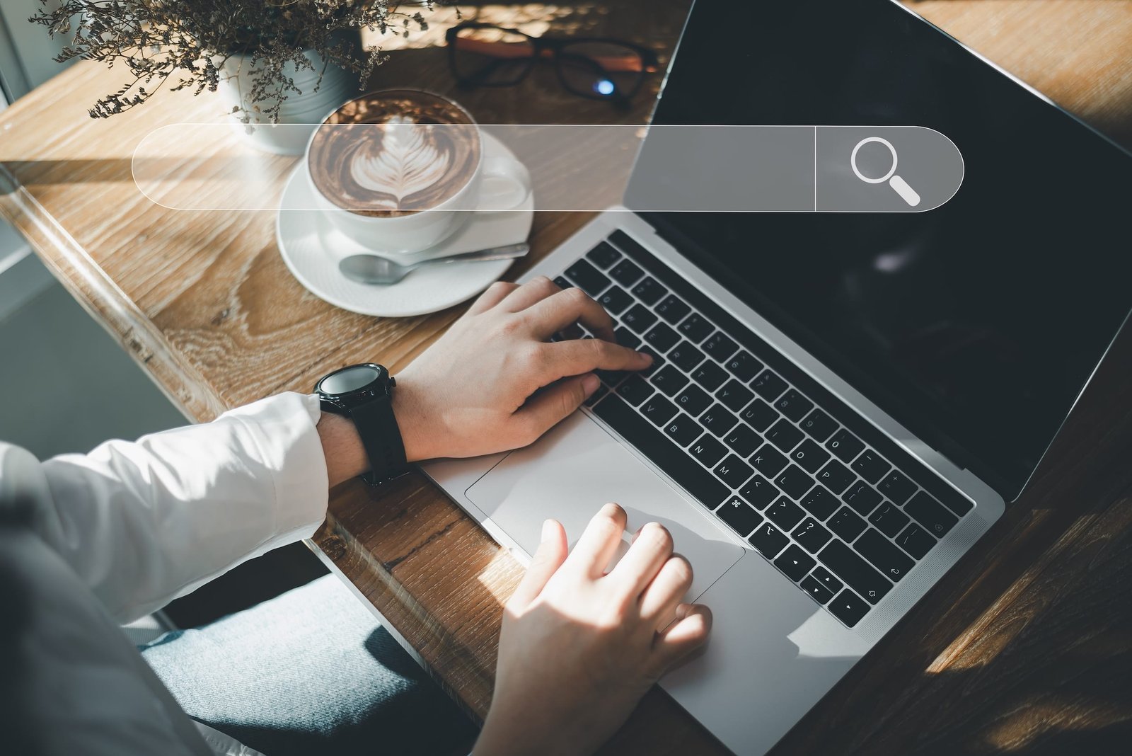 Person typing on a laptop at a wooden table with a cup of coffee, sunglasses, and plant nearby; a digital search bar graphic is overlaid on the image.