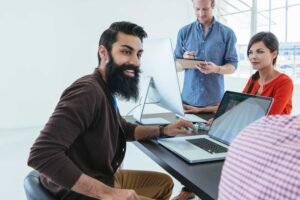 Four people work together in a bright office; one man sits at a desk using a laptop and smiles at the camera while the others collaborate on an audit in the background.
