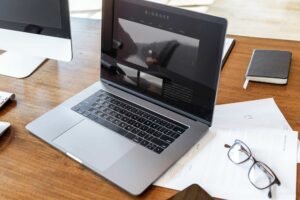 A laptop displaying a website is on a wooden desk with papers, eyeglasses, a pen, and a closed notebook nearby.