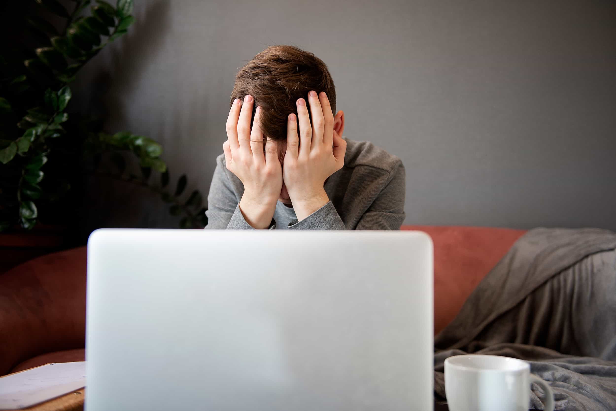 A person sits on a couch behind an open laptop, covering their face with both hands. A coffee mug is on the table in front of them.