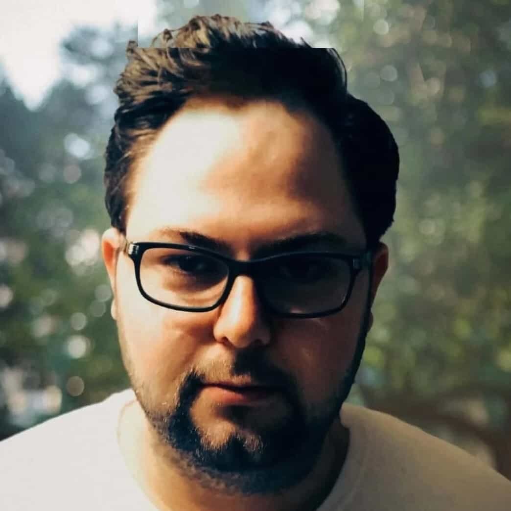 Free Consultation A man with short dark hair, glasses, and facial hair is looking at the camera as if ready for a consultation. He is wearing a light-colored shirt with a blurred outdoor background.
