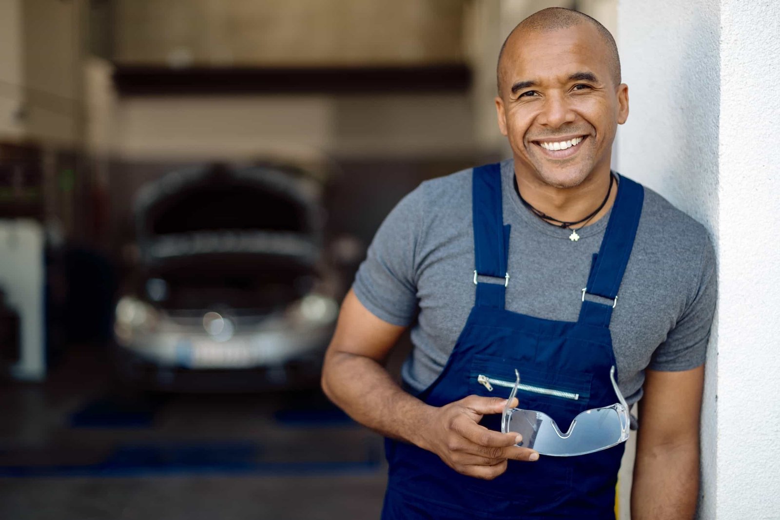Smiling mechanic in blue overalls holding safety goggles stands in a garage with a car in the background.