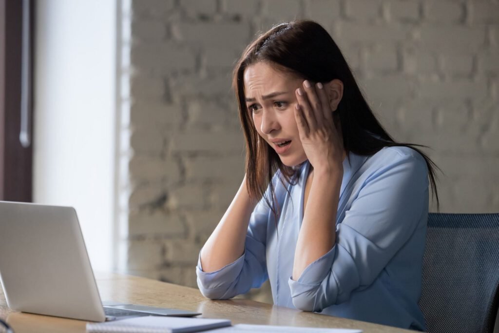 A woman sits at a desk looking at a laptop with a worried expression, holding her head with both hands. The Hidden Costs of Using Wix and Squarespace for Your Business