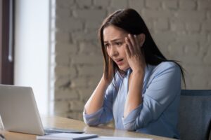 A woman sits at a desk looking at a laptop with a worried expression, holding her head with both hands. The Hidden Costs of Using Wix and Squarespace for Your Business