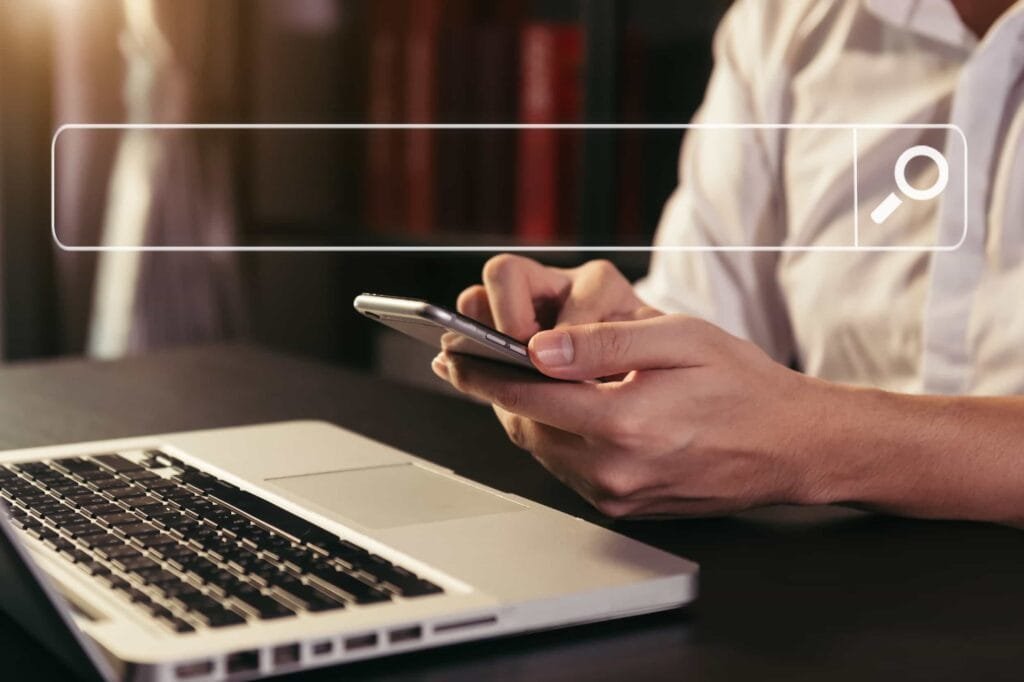 Why isn’t my old website showing up on Google? A person in a white shirt uses a smartphone next to an open laptop, with a digital search bar graphic overlay at the top of the image.