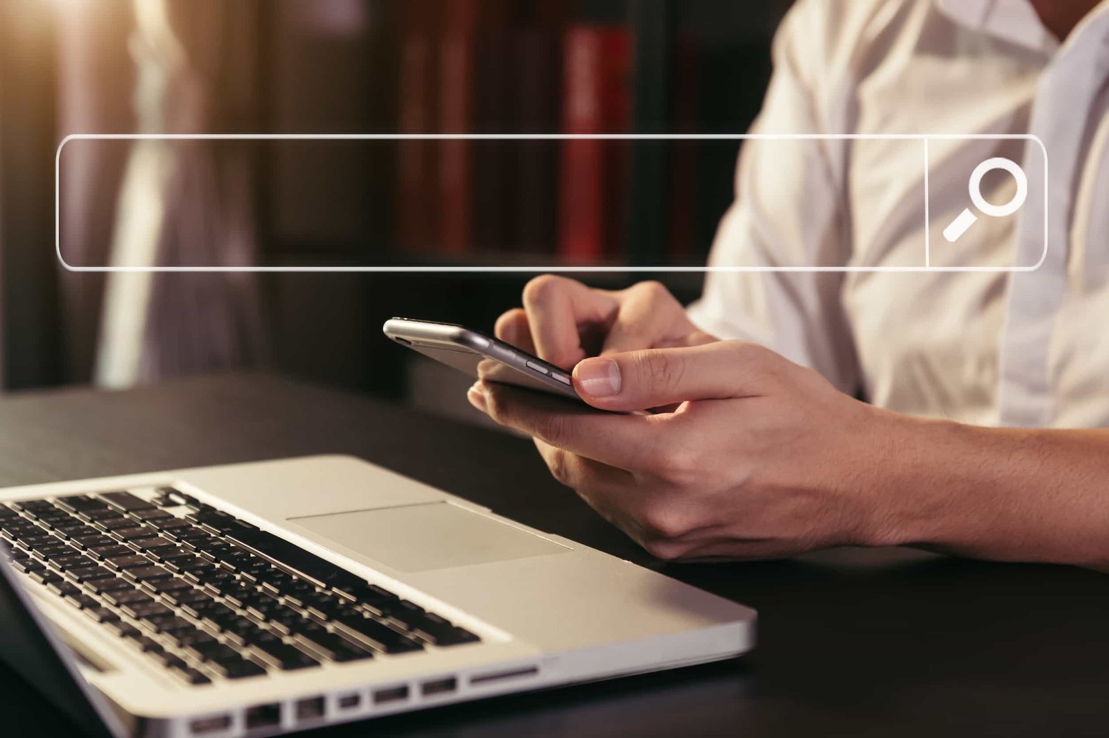 A person in a white shirt uses a smartphone next to an open laptop, with a digital search bar graphic overlay at the top of the image.
