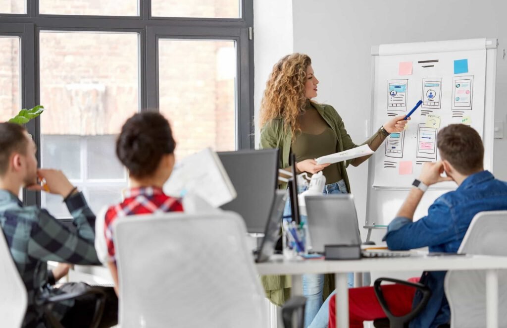 A woman presents mobile app wireframes on a flip chart to three seated colleagues in a modern office setting, sparking ideas on how to refresh my website.