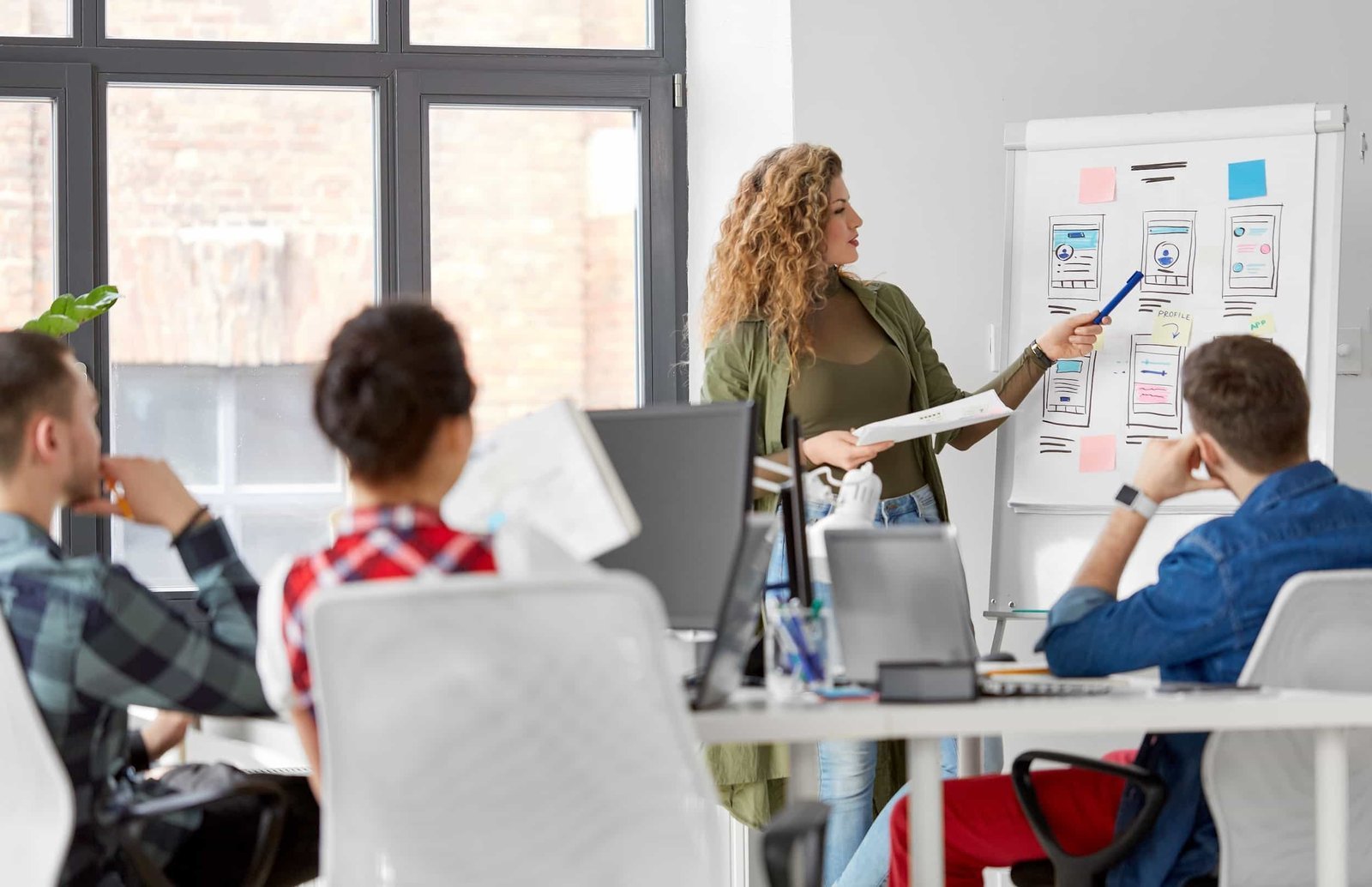 A woman presents mobile app wireframes on a flip chart to three seated colleagues in a modern office setting, sparking ideas on how to refresh my website.