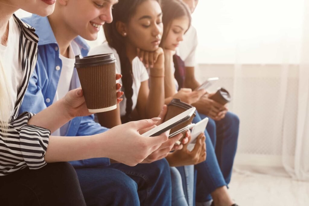 Five young people sitting in a row, holding coffee cups and looking at their smartphones and tablets. Search Intent Changes in SEO.