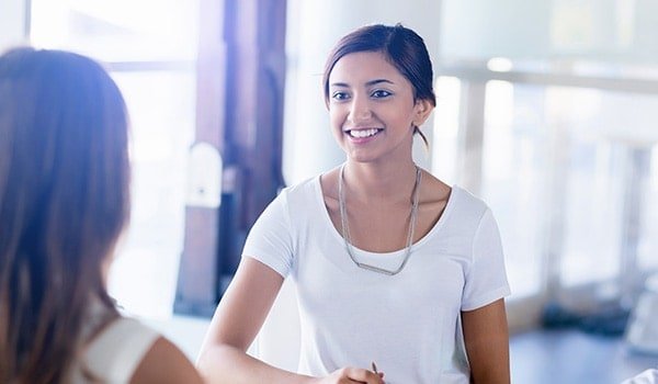 A woman in a white shirt smiles and shakes hands with another person in a bright, indoor setting.