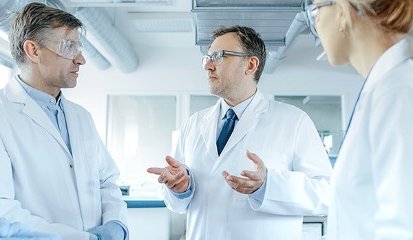 Three scientists in lab coats and safety glasses have a discussion in a laboratory setting.