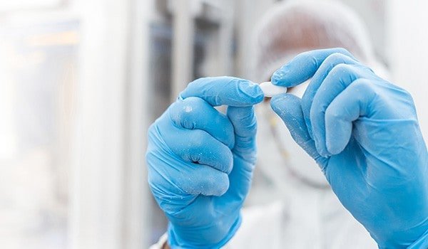 A person wearing blue gloves and protective gear holds a white pill or tablet between their fingers in a laboratory setting.