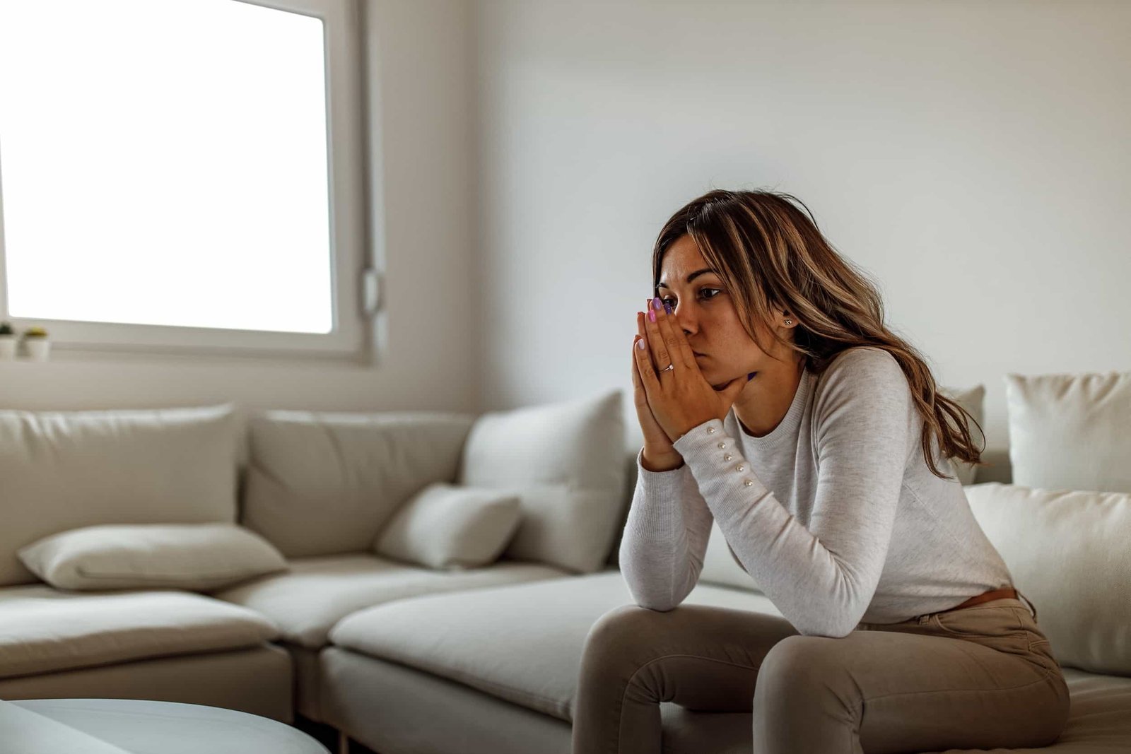 A woman sits on a couch with her hands covering her mouth, appearing deep in thought or concerned, in a bright living room. Traffic