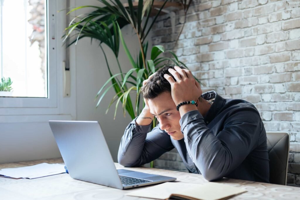 A person sits at a table holding their head in frustration while looking at a laptop, with papers and a notebook nearby.