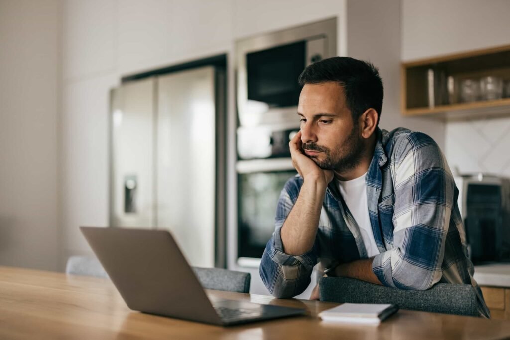 A man sits at a kitchen table, resting his face on his hand while looking at an open laptop with a notepad and pen nearby.