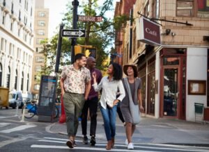 Four people walk together across a city street crosswalk, smiling and talking, with buildings, street signs, and a café in the background. Queens NY Web Design