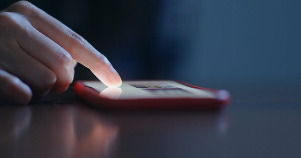 A close-up of a person’s finger scrolling on the screen of a smartphone with a red case, resting on a dark surface.