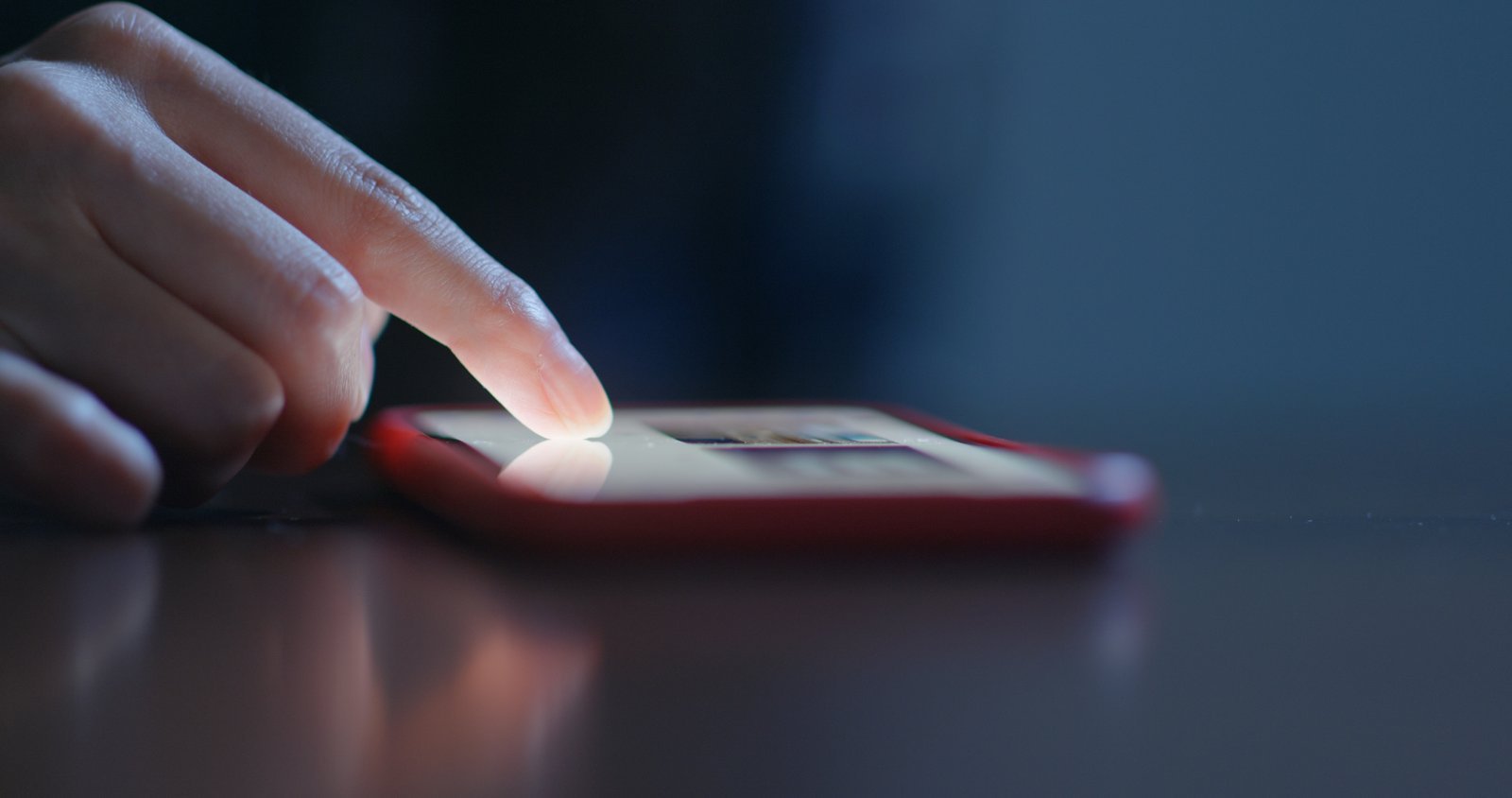 A close-up of a person’s finger scrolling on the screen of a smartphone with a red case, resting on a dark surface.