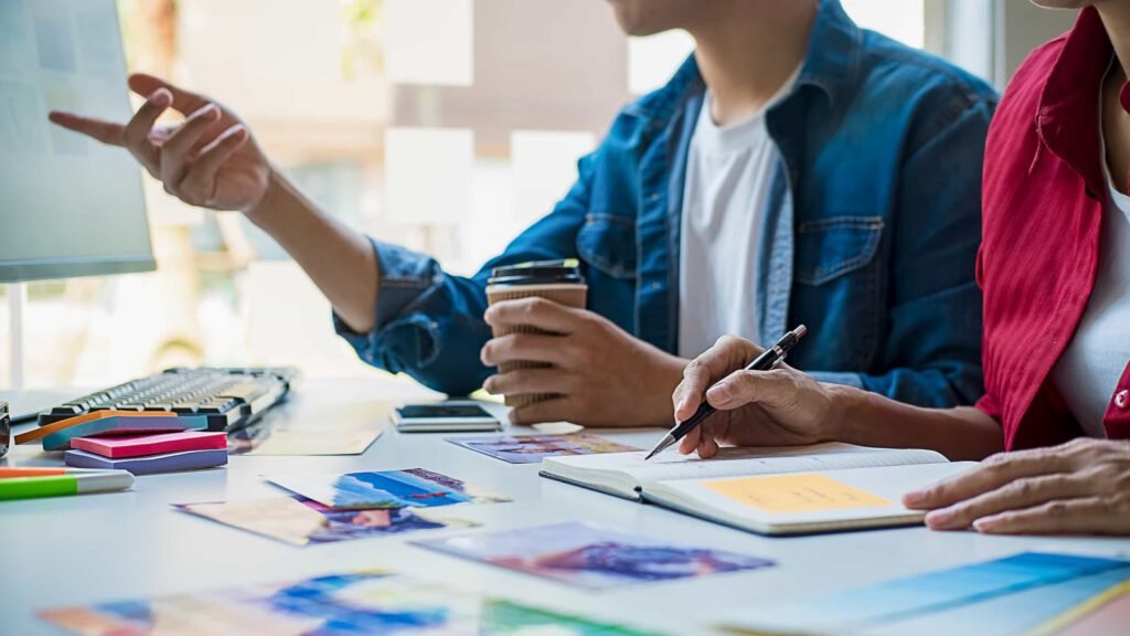 Two people sit at a desk with photos and stationery, one taking notes in a notebook, the other pointing at a monitor and holding a coffee cup.