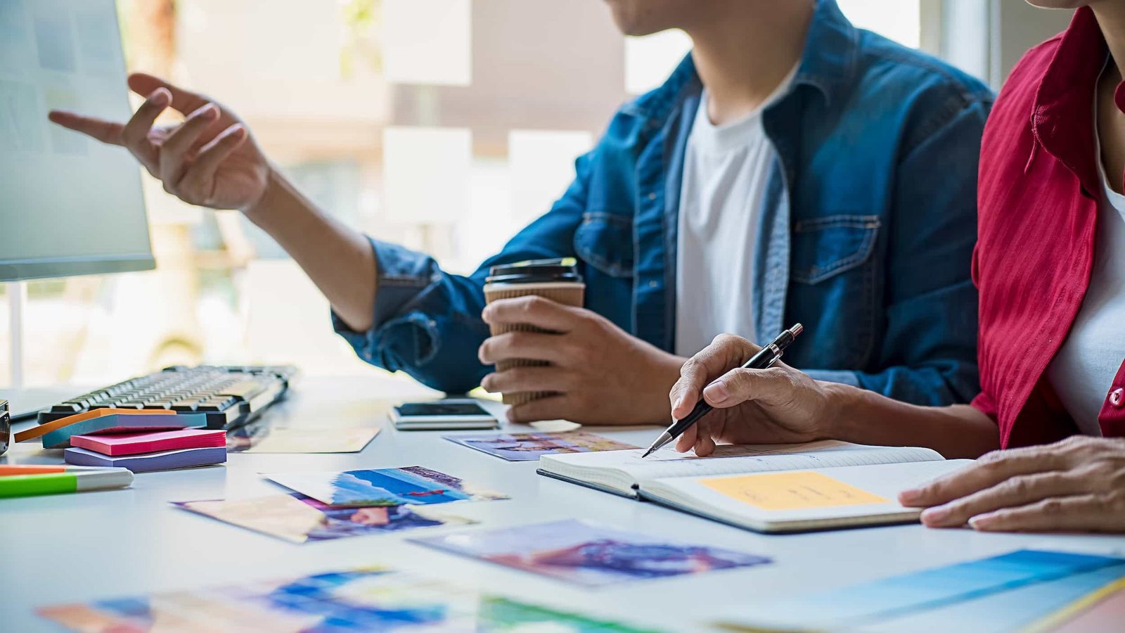 Two people sit at a desk with photos and stationery, one taking notes in a notebook, the other pointing at a monitor and holding a coffee cup.