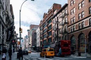 City street with brick buildings, pedestrians on the sidewalk, yellow taxis, a red double-decker bus, and medical shops visible on a cloudy day. nyc medical