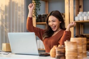 A woman sits at a desk with wooden items, smiling and raising her fists in excitement while looking at a laptop, embodying the spirit of "I May Not Be an Agency, But Here's What I Do Better. Agency