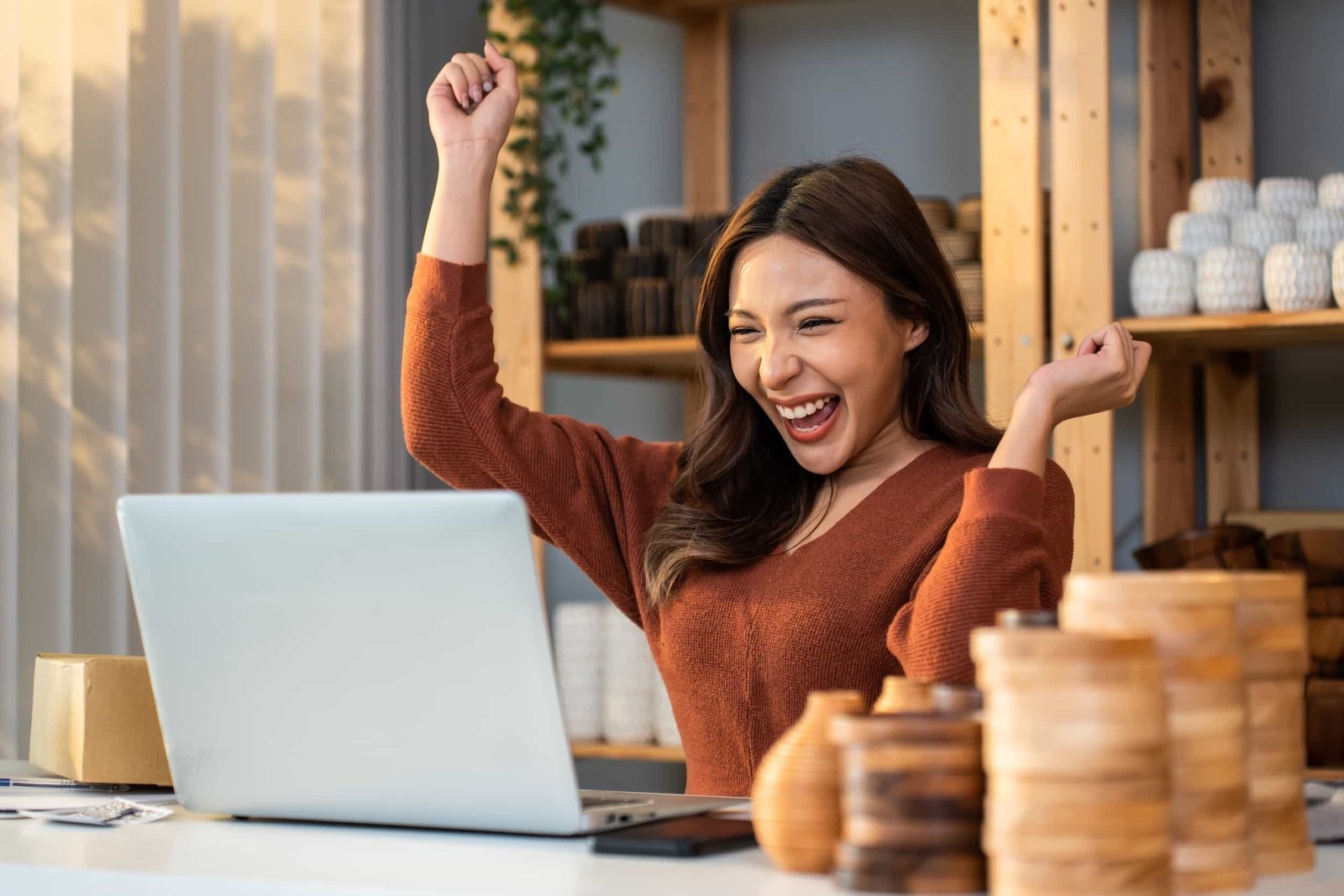 A woman sits at a desk with wooden items, smiling and raising her fists in excitement while looking at a laptop, embodying the spirit of "I May Not Be an Agency, But Here's What I Do Better. Agency