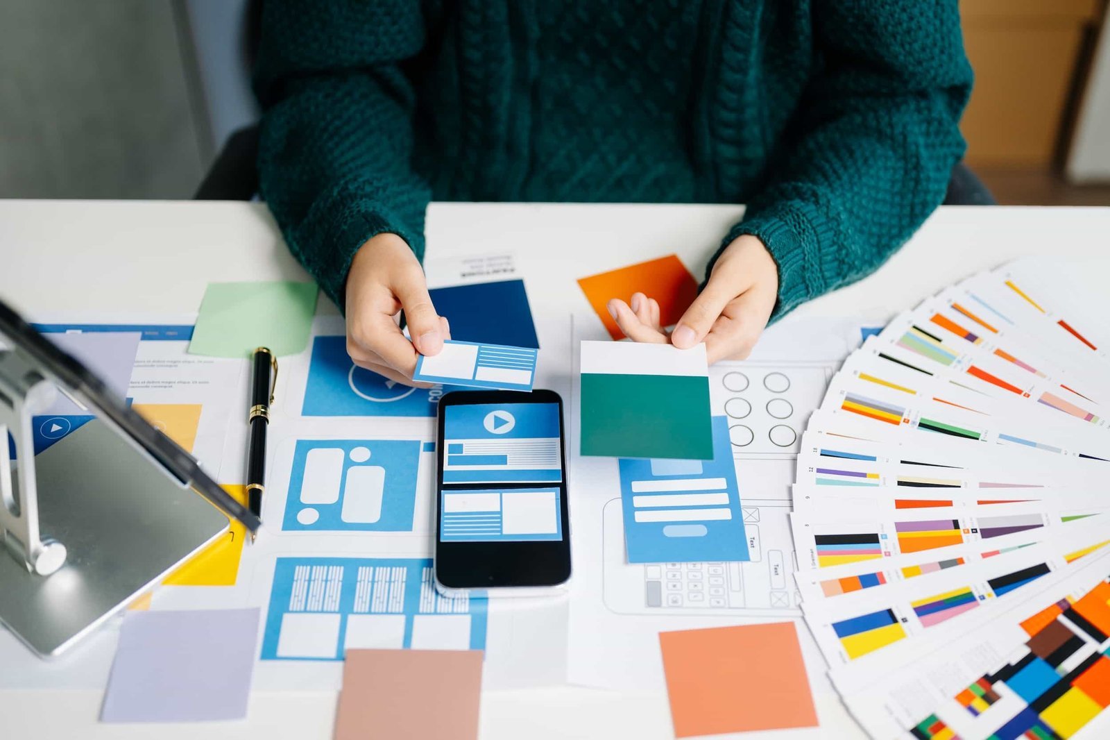 Person selecting color swatches at a desk with a smartphone, color samples, web design layouts, and a color palette fan spread out.