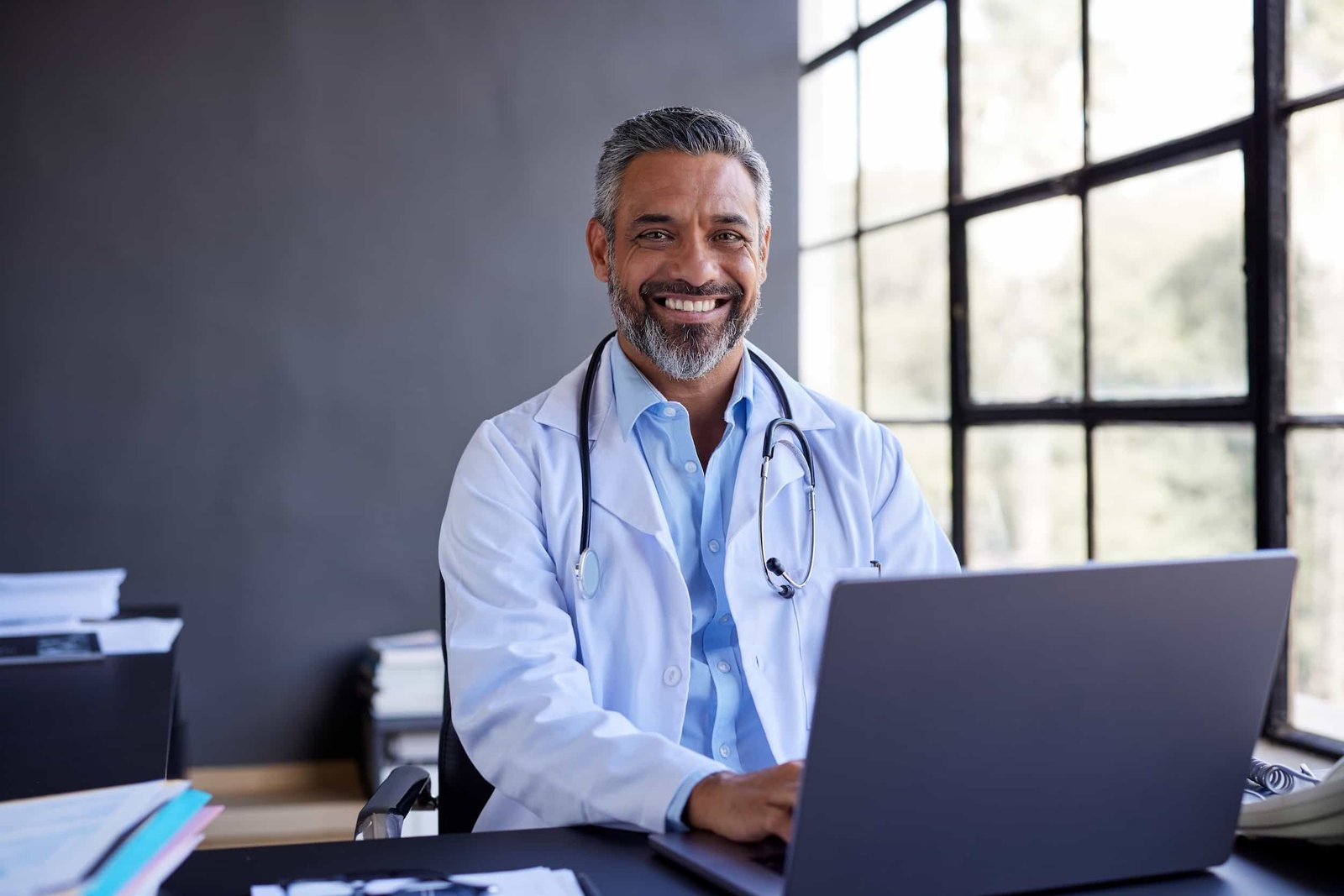 A man in a white lab coat with a stethoscope around his neck sits at a desk, smiling at the camera while using a laptop in a bright office. NYC patients decide in 4 seconds if they'll book with you. I audit medical sites weekly. Here are the 5 signals most practices in Queens and Manhattan miss.