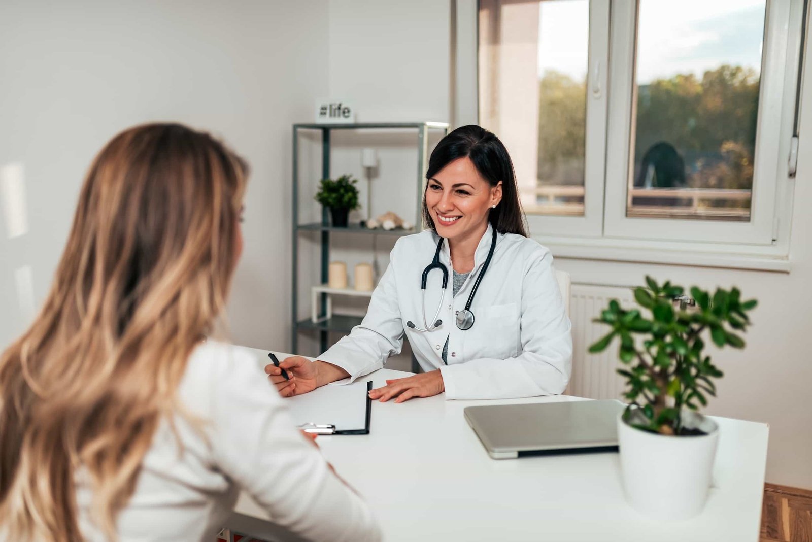 A female doctor in a white coat with a stethoscope smiles while speaking with a patient at her medical office in Queens, NY. SEO