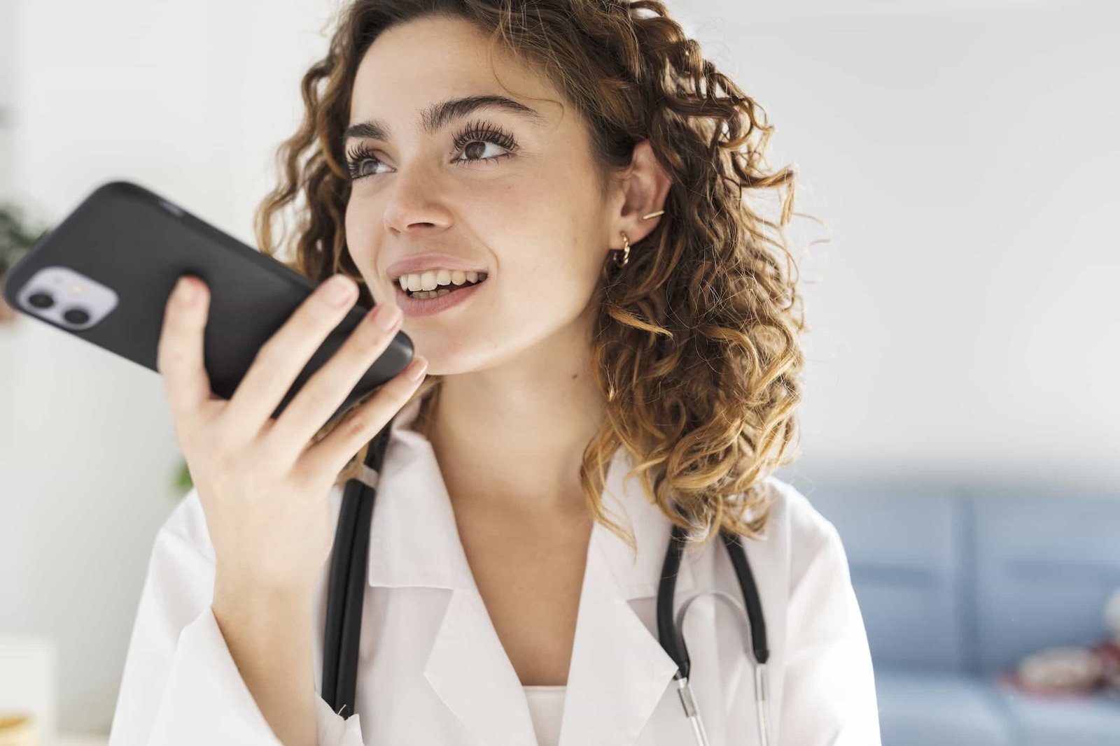 A woman wearing a white coat and stethoscope speaks into a smartphone, appearing to use a voice message or virtual assistant in a bright indoor setting. The Instagram Audit Every Doctor Should Run.