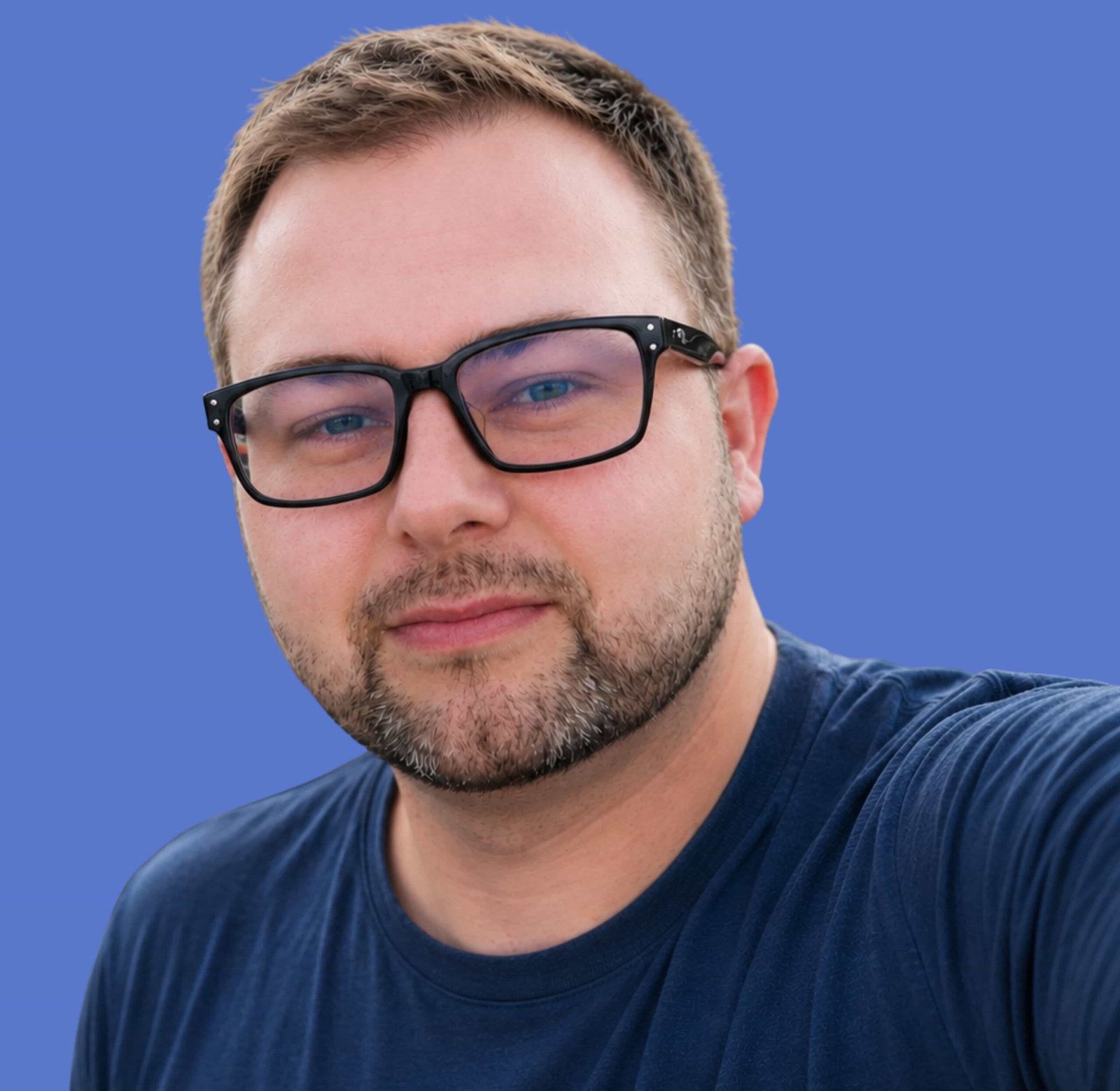 A man with short brown hair, a beard, and glasses wearing a navy blue medical t-shirt stands in front of a solid blue background.
